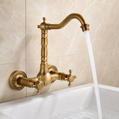 An antique brass wall-mounted faucet with a curved spout and two cross handles, shown installed above a white sink with water running from the spout against a beige tiled wall.